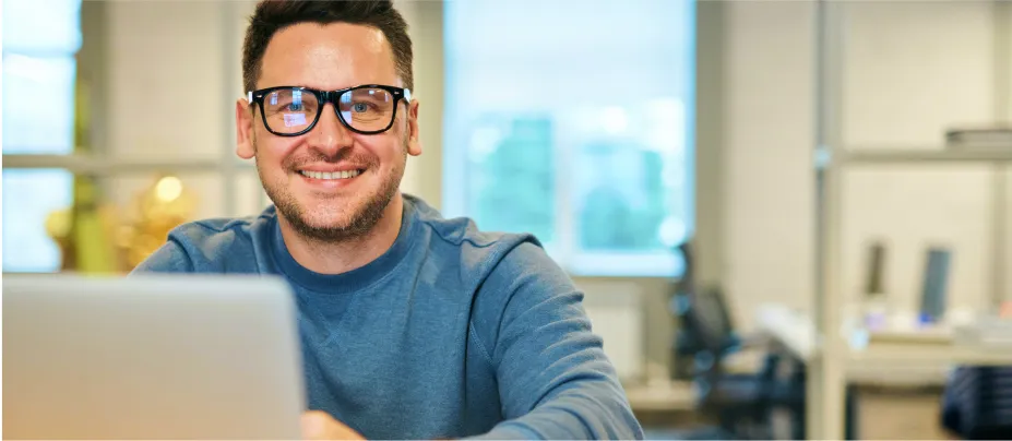 Un señor con lentes en su oficina sonriendo detrás de su computadora